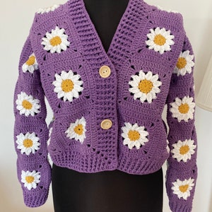 Könnte beinhalten: Eine lilafarbene Häkeljacke mit weißen Gänseblümchen. Die Jacke hat einen V-Ausschnitt und einen Knopfverschluss.