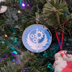 May include: A Christmas tree ornament featuring a blue and white Fallout logo with a red ribbon and a Santa Claus ornament hanging below.