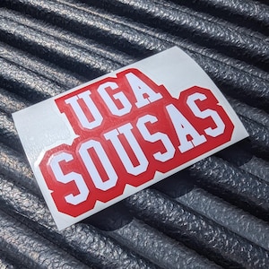 May include: Red and white vinyl sticker with the text "UGA SOUSAS" in a bold, blocky font.