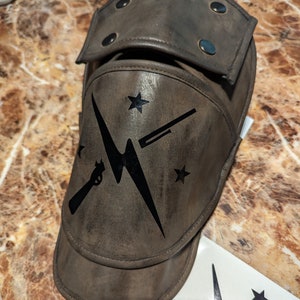 Può includere: Un ginocchiere in pelle marrone con un design a forma di fulmine nero e due fucili incrociati. Il ginocchiere ha una patta che può essere ripiegata sulla parte superiore. Il design include anche due stelle.