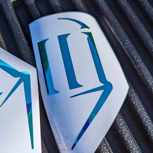 May include: A blue and green iridescent vinyl decal with the letter "R" in a stylized font. The decal is on a white sheet of transfer paper with a grid pattern.
