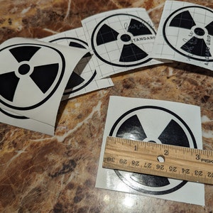 May include: Four black and white radiation warning stickers on a brown surface. The stickers are approximately 2 inches in diameter. The text on one sticker reads "lauron STANDARD". The text on another sticker reads "ADADE GRIP".