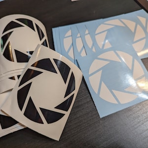 May include: A stack of white stickers with a black aperture symbol, and a sheet of blue stickers with a white aperture symbol.