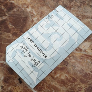 May include: A white vinyl stencil with the word "Love" in a decorative font. The stencil is on a blue sheet with a grid pattern and the text "Standard Grip,  Love of Life Basics" printed on it.