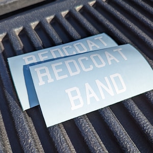 May include: Two white vinyl stickers with the text "REDCOAT BAND" on a blue background. The stickers are on a black textured surface.