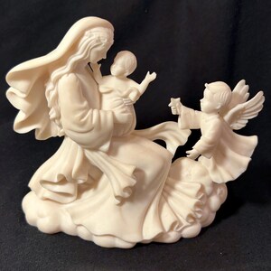 May include: A white porcelain figurine of the Virgin Mary holding a baby Jesus. A small angel with wings is reaching out to the baby.