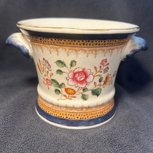 May include: A white ceramic planter with two handles, decorated with a floral pattern in shades of pink, green, blue, and orange. The planter has a gold trim around the top and bottom.