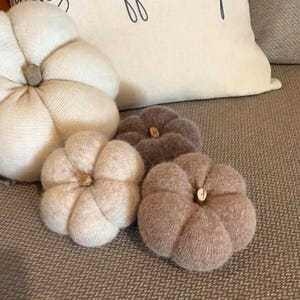 May include: A selection of fabric pumpkins in shades of cream, beige, and brown. The pumpkins have a textured appearance, possibly knitted or felted. The background includes a cream cushion with black script. The pumpkins are ideal for autumn decor.