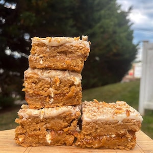 May include: A stack of five square-shaped baked goods with a light-colored frosting and a sprinkle of nuts on top. The baked goods are stacked on a wooden cutting board, with a blurred green background.
