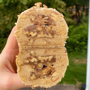 Könnte beinhalten: Nahaufnahme eines in der Mitte durchgeschnittenen Pekannuss-Pralinen-Kekses, der einen hellbraunen Teig enthüllt, der mit gehackten Pekannüssen gefüllt ist. Der Keks ist mit einer dünnen Schicht weißen Zuckergusses bedeckt.