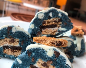 May include: Close-up of blue cookie dough balls with white chocolate and chocolate chips, layered with cookie wafers and topped with white chocolate. The cookie dough balls are cut in half to show the layers.