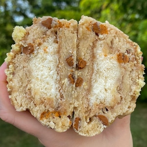 以下が含まれることがあります： 半分にカットされたクッキーのクローズアップ。クリーミーな白いフィリングとチョコレートチップが露出しています。クッキーは薄い茶色で、少しざらざらした食感です。