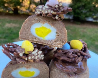 Puede incluir: Tres nidos de huevos de Pascua de chocolate con un huevo de caramelo amarillo dentro de cada uno. Los nidos están cubiertos con pretzels cubiertos de chocolate y huevos de caramelo.