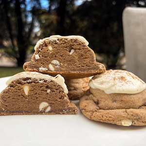 Op de afbeelding: Drie bruine koekjes op een wit bord. Twee koekjes zijn doormidden gesneden, wat een dichte, textuurrijke binnenkant met zichtbare witte chocoladestukjes onthult. Het derde koekje is heel, bedekt met wit glazuur en een snufje bruine kruiden.
