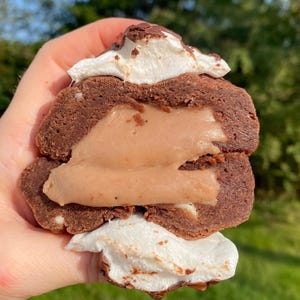 May include: A chocolate brownie cookie with a layer of marshmallow and a creamy filling. The cookie is held in a hand.