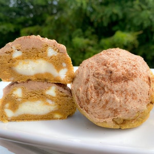 Peut inclure: Deux biscuits à la citrouille et aux épices avec des pépites de chocolat blanc et une garniture de fromage à la crème. Un biscuit est coupé en deux, révélant la garniture. Les biscuits sont sur une assiette blanche.
