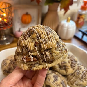 Op de afbeelding: Een rond, goudbruin koekje met zichtbare pecannotenstukjes en een witte glazuurlaag. Het koekje heeft een textuurrijk, kruimelig oppervlak en wordt getoond voor andere soortgelijke koekjes in een witte rechthoekige schaal.