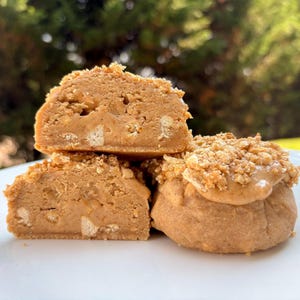 May include: Close-up of three pieces of peanut butter fudge. The fudge is light brown with a crumbly topping and visible biscuit pieces. One piece is cut to reveal the interior. The fudge is stacked on a white plate.