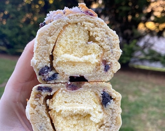 Puede incluir: Primer plano de un pastel relleno de crema blanca y arándanos. El pastel está cortado por la mitad y el relleno es visible. El pastel está cubierto con un glaseado blanco.