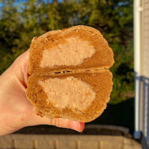 May include: A close-up of a pumpkin spice cookie cut in half, revealing a pink cream cheese filling. The cookie is glazed with a light brown glaze.