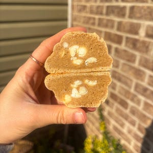 May include: A close-up of a cookie cut in half, revealing a soft, light brown interior with white chocolate chips. The cookie is held in a hand, with a blurred background of a brick wall and greenery.