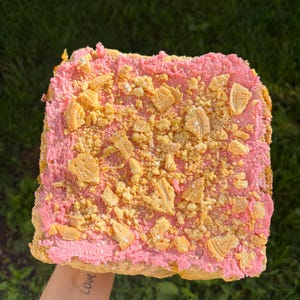 May include: A square dessert with pink frosting and a topping of golden-brown, irregularly shaped pieces. The dessert is held against a backdrop of green grass. The words "I Love" are tattooed on the person's arm holding the dessert.
