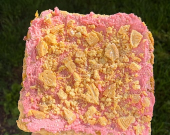 Puede incluir: Un postre cuadrado con glaseado rosa y una cobertura de trozos dorados irregulares. El postre se sostiene sobre un fondo de hierba verde. Las palabras "I Love" están tatuadas en el brazo de la persona que sostiene el postre.