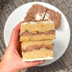 Puede incluir: Dos rebanadas de pastel en capas con una base marrón claro, un relleno marrón oscuro y un borde de glaseado blanco. El pastel se sostiene en una mano, con una rebanada de pastel más grande en el fondo sobre un plato blanco. El pastel tiene glaseado de chocolate.