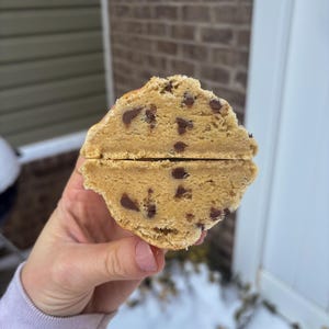 Puede incluir: Primer plano de una galleta con chispas de chocolate cortada por la mitad, revelando el interior. La galleta es de color marrón claro con chispas de chocolate visibles. El corte muestra un centro suave y pastoso, lo que sugiere un dulce fresco y casero. La galleta se sostiene en una mano.