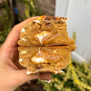May include: A thick, golden-brown cookie broken in half to reveal a soft, gooey centre filled with melted marshmallow and caramel. The cookie dough is studded with small chocolate pieces and bits of toasted marshmallow on the top surface.