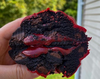 May include: A close-up of a chocolate chip cookie with red velvet frosting. The cookie is broken in half, revealing the layers of frosting and chocolate chips.