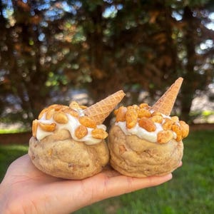 May include: Two round, textured, light brown cookie dough balls topped with white icing, golden roasted peanuts, and a small, pointed waffle cone piece inserted into the centre of each, creating a miniature ice cream cone appearance.