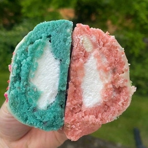 Puede incluir: Primer plano de un postre cortado por la mitad, que revela un núcleo de malvavisco blanco dentro de un pastel azul y rosa. El pastel tiene una superficie texturizada y está decorado con chispas. Los colores son vibrantes.