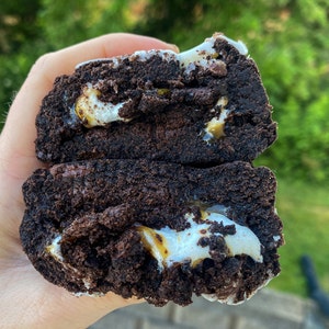 May include: A close-up of a chocolate brownie cookie with marshmallow and caramel filling. The cookie is broken in half, revealing the gooey filling inside.