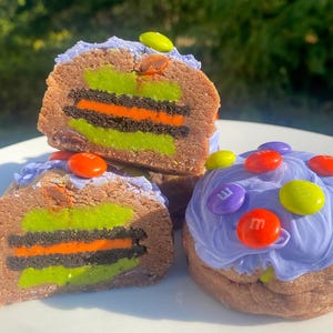 Peut inclure: Trois boules de pâte à biscuits sur le thème d'Halloween. L'une est coupée en deux pour révéler des couches de glaçage vert et orange, un biscuit Oreo noir et une couche verte. Les deux autres sont recouvertes de glaçage violet et décorées de M&Ms verts et orange.