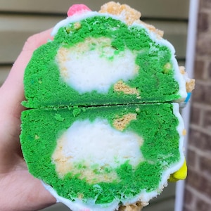 May include: A cross-section of a green cake roll with a white filling. The cake has a vibrant green exterior and a white, creamy interior. The top is decorated with sprinkles and small pieces of a light-colored crumble.