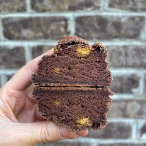 Puede incluir: Un brownie de chocolate cortado por la mitad con trozos visibles de galletas doradas. El brownie tiene un color marrón oscuro intenso y una superficie ligeramente texturizada. La imagen destaca el interior húmedo y las texturas contrastantes.