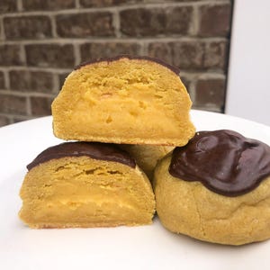 May include: A stack of three golden-yellow pastries with a creamy filling and a dark chocolate topping. One pastry is whole, while the others are cut in half, revealing the interior. The pastries are arranged on a white plate.