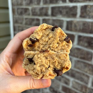 以下が含まれることがあります： チョコレートチップクッキーを半分に切ったクローズアップ。柔らかく、質感のある内部にダークチョコレートチップが見えます。クッキーはゴールデンブラウンで、少し崩れやすい外観です。背景はレンガの壁です。