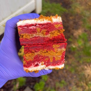 May include: A layered dessert with red, tan, and white layers. The top layer is white with a crumbly topping. The red layers appear to be a cake or brownie, and the tan layers have a textured appearance. The dessert is cut in half.