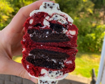 May include: A red velvet cookie with a layer of chocolate cookie and white chocolate chips. The cookie is held in a hand.