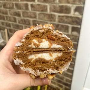 Op de afbeelding: Doorsnede van een dessert met een witte vulling, omgeven door een bruine, cake-achtige buitenkant. De boven- en onderranden zijn bedekt met een witte glazuur en gehakte pecannoten. Het dessert wordt in een hand gehouden tegen een bakstenen achtergrond.