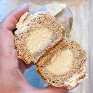 May include: A close-up of a baked pastry cut in half, revealing a creamy, light-colored filling. The pastry has a layered, textured appearance with a light brown exterior. The background includes a piece of the same pastry and a wooden surface.