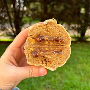 May include: A round, golden-brown baked biscuit broken in half to reveal a soft, textured interior with a visible layer of dark, sticky caramel-like filling containing small nut pieces embedded in the centre of the dough.
