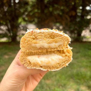 May include: A golden-brown biscuit broken in half to reveal a thick, creamy white filling. The biscuit has a textured, crumbly exterior with small bits of topping and a soft, dense interior surrounding the smooth, pale centre layer.