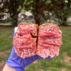 Op de afbeelding: Een close-up van een roze glazuurgebakje dat doormidden is gesneden en een roze vulling onthult. Het gebakje wordt vastgehouden door een gehandschoende hand. Het gebakje heeft witte glazuur en hagelslag bovenop. Het woord "Love You" is op de arm van de persoon getatoeëerd.