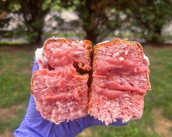 May include: A close-up of a pink frosted pastry cut in half, revealing a pink filling. The pastry is held by a gloved hand. The pastry has white frosting and sprinkles on top. The word "Love You" is tattooed on the person's arm.