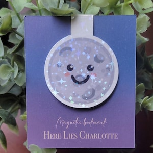 Może przedstawiać: Fioletowo-niebieskie tło z gradientami z białym, księżycowym magnesem do zakładek. Księżyc ma uroczą kreskówkową twarz z uśmiechem i błyszczącymi akcentami z brokatu. Na zakładce nadrukowany jest tekst "Magnetic Bookmark" i "HERE LIES CHARLOTTE".