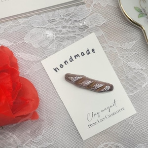 May include: Handmade clay magnet shaped like a baguette, with the text "Clay magnet" and "Here Lies Charlotte" on a white card with the word "handmade" written on it.