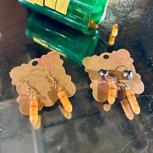 May include: Earrings with orange capsule-shaped charms, each labeled "Amphet 20 mg", dangle from silver hooks. The earrings are displayed on brown cardstock. A green pill bottle and loose capsules are also visible.